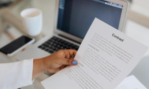 A hand holding a contract over a laptop on a desk.