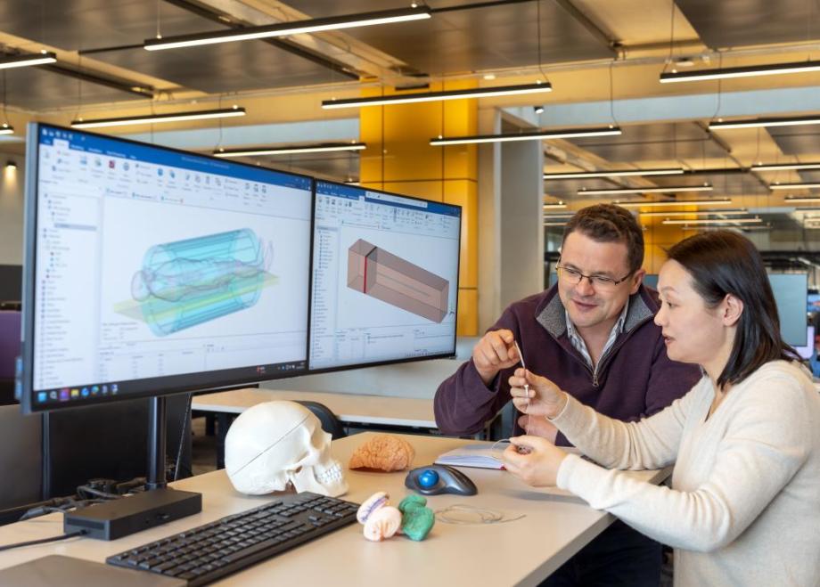 Members of TTP's Neurotechnology team look closely at a MRI-safe implant, holding it up to see it better. The individuals are around a desk, and on the screen is some computer modelling of an individual within an MRI machine.