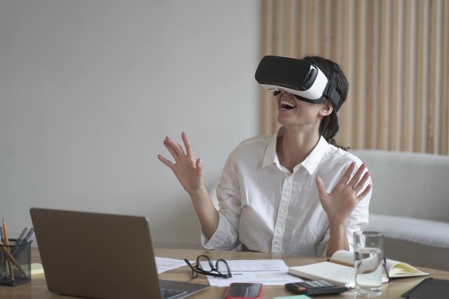 Business lady sits at work desk wearing a VR headset for laptop, raising her hands up as if trying to touch objects in 3D reality
