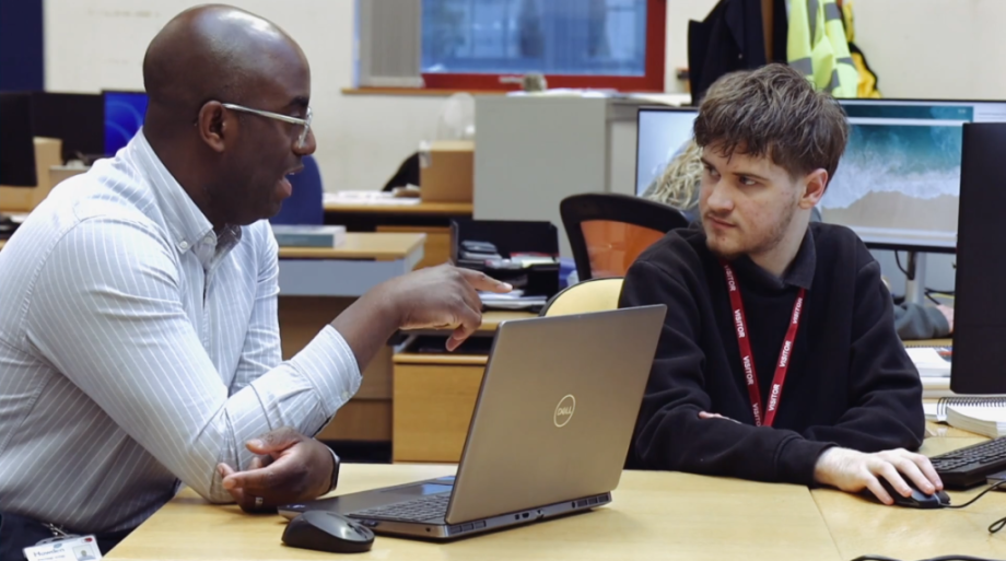 A man with glasses and a striped shirt points toward a laptop screen while speaking to a young man wearing a "VISITOR" lanyard in an office setting.