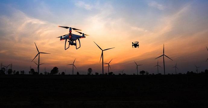 Drones fly above a field with wind turbines in it at sunset