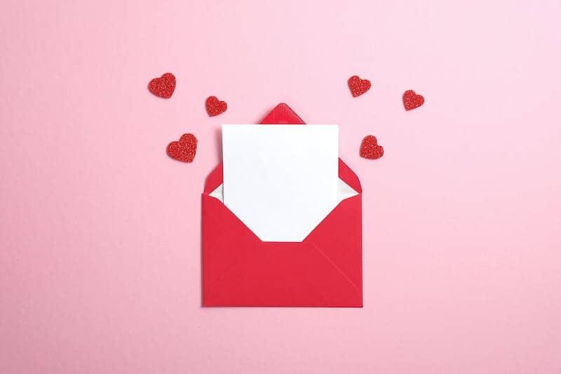 Red envelope with white card and love hearts emerging, sitting on a pink background