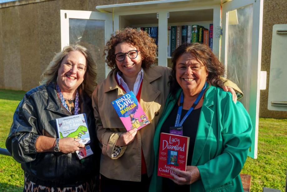 Three women stood in front of Little Free Library holding books