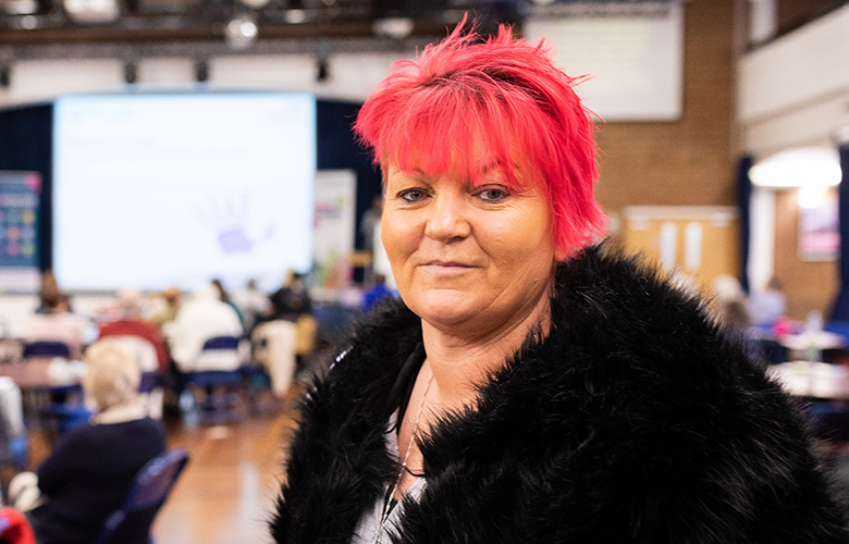 Red haired woman wearing a black coat, indoors at a Healthwatch event is looking at the camera. 